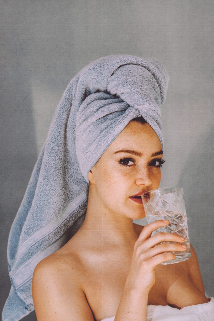 A woman wearing white robe, hair wrapped in towel. She is drinking water from a transparent glass while her eyes are fixed at the camera.