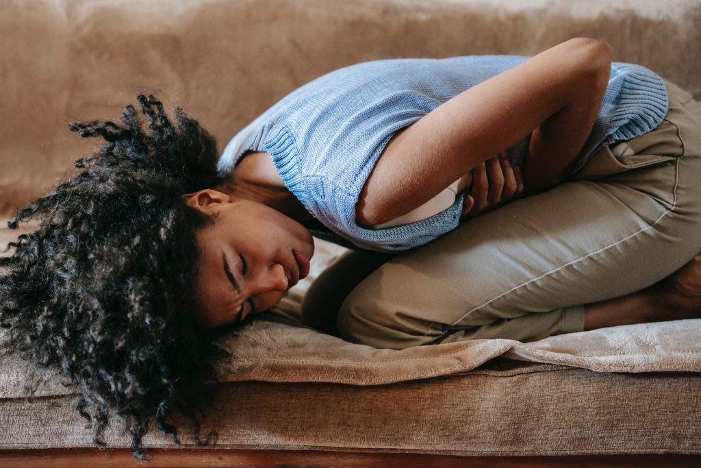 A black woman in blue sleeveless shirt, curling her body, as if crouching in pain form bloating or constipation.